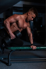 Fototapeta na wymiar Strong athletic young man with naked torso and muscles doing dumbbell workout in the gym at night