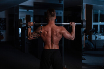 Fototapeta na wymiar Sports man trainer with muscular back and tattoo on his shoulder doing exercises with dumbbells in the gym on a blue dark background