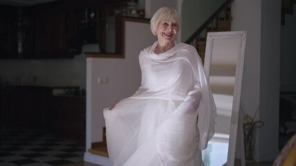 Slim beautiful senior bride spinning in white elegant wedding dress in slow motion indoors. Portrait of happy charming Caucasian woman posing in living room at home on wedding day smiling