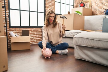 Young woman holding piggy bank and hammer moving to a new home clueless and confused expression....