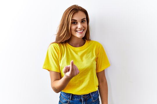 Young brunette woman standing over isolated background beckoning come here gesture with hand inviting welcoming happy and smiling