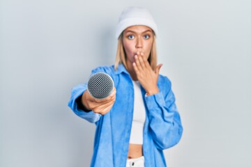 Beautiful blonde woman holding reporter microphone to the camera covering mouth with hand, shocked...