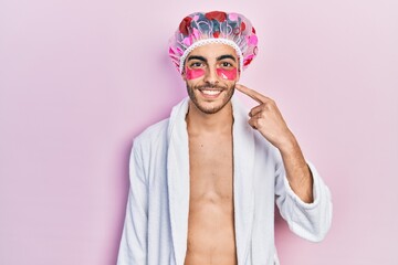 Young hispanic man wearing bathrobe and shower cap using eyes bags patches smiling happy pointing with hand and finger