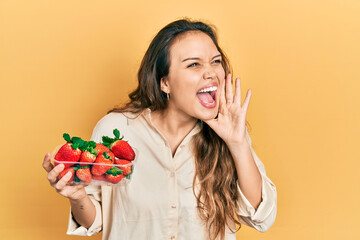 Young hispanic girl holding strawberries shouting and screaming loud to side with hand on mouth. communication concept.