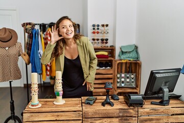 Beautiful hispanic woman working at fashion shop smiling with hand over ear listening an hearing to rumor or gossip. deafness concept.