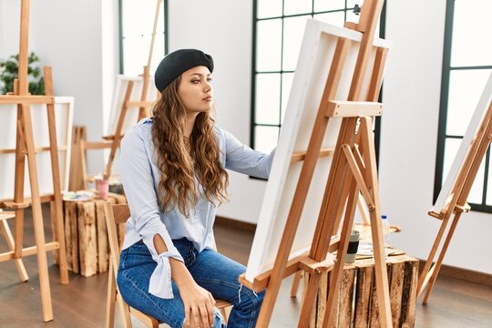 Young hispanic artist woman painting on canvas at art studio looking to side, relax profile pose with natural face and confident smile.