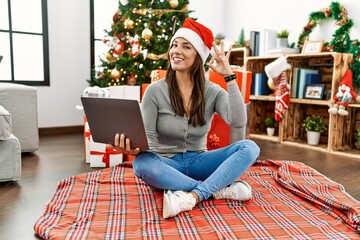 Young latin woman using laptop sitting by christmas tree smiling positive doing ok sign with hand and fingers. successful expression.