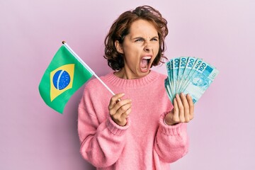 Young brunette woman holding brazil flag and real banknotes angry and mad screaming frustrated and...