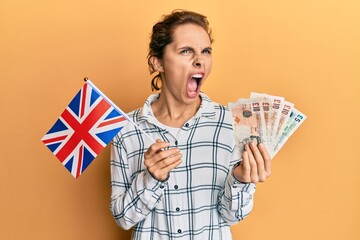 Young brunette woman holding uk flag and pounds banknotes angry and mad screaming frustrated and...