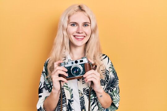Beautiful caucasian woman with blond hair holding vintage camera smiling with a happy and cool smile on face. showing teeth.