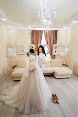 bride in white coat by wedding dress in room of her house. sofa with balloons.