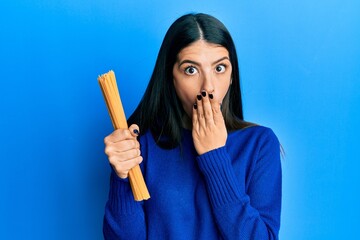 Young hispanic woman holding uncooked spaghetti covering mouth with hand, shocked and afraid for...