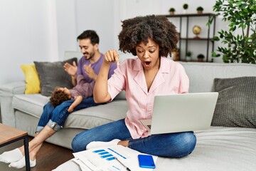 Mother of interracial family working using computer laptop at home pointing down with fingers showing advertisement, surprised face and open mouth