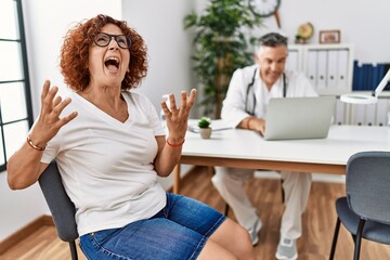 Senior woman sitting at doctor appointment crazy and mad shouting and yelling with aggressive expression and arms raised. frustration concept.