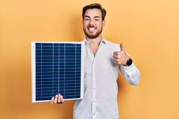 Handsome caucasian man with beard holding photovoltaic solar panel smiling happy and positive,...