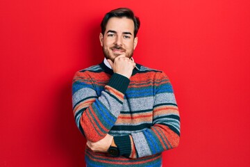 Handsome caucasian man with beard wearing elegant wool winter sweater looking confident at the camera smiling with crossed arms and hand raised on chin. thinking positive.