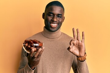 Young african american man holding bowl with dates doing ok sign with fingers, smiling friendly gesturing excellent symbol