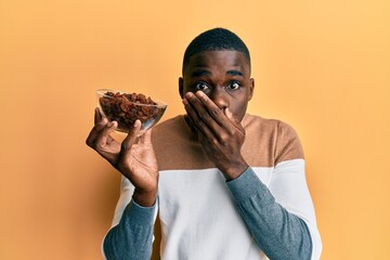 Young african american man holding bowl with raisins covering mouth with hand, shocked and afraid...