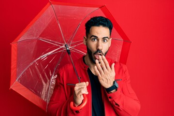 Young hispanic man holding umbrella covering mouth with hand, shocked and afraid for mistake....