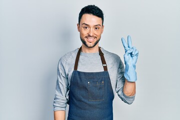 Young hispanic man wearing apron and gloves smiling with happy face winking at the camera doing...