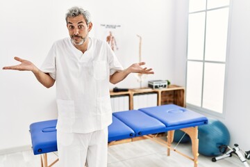 Obraz premium Middle age hispanic therapist man working at pain recovery clinic clueless and confused expression with arms and hands raised. doubt concept.