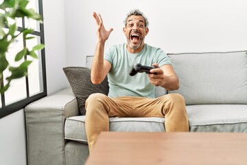 Middle age hispanic man playing video game sitting on the sofa celebrating victory with happy smile and winner expression with raised hands