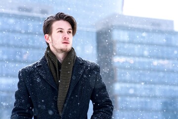 Portrait of handsome serious freezing frozen man, young blonde European guy in coat and scarf, office worker is going to job, work in snow at winter snowy cold frosty day. Modern building background