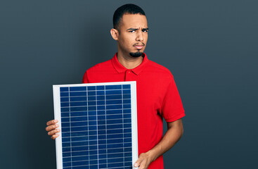 Young african american man holding photovoltaic solar panel clueless and confused expression. doubt...