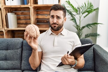 Handsome hispanic man holding brain working at psychology clinic skeptic and nervous, frowning...