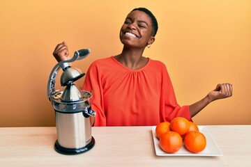 Young african american woman sitting on the table using juicer very happy and excited doing winner...