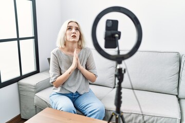 Fototapeta na wymiar Young caucasian woman recording vlog tutorial with smartphone at home begging and praying with hands together with hope expression on face very emotional and worried. begging.