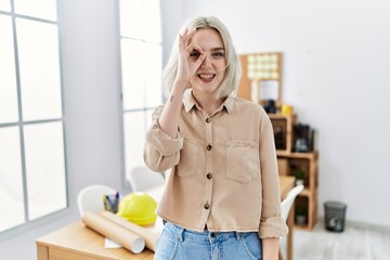 Young beautiful caucasian woman at construction office doing ok gesture with hand smiling, eye looking through fingers with happy face.