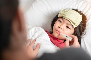 Asian mother measuring temperature girl with digital thermometer in her mouth on bed at morning time, Sick child have cool towel for reduce high fever,  Selective focus, Healthy and infection concept