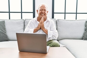 Senior man using laptop at home sitting on the sofa begging and praying with hands together with...