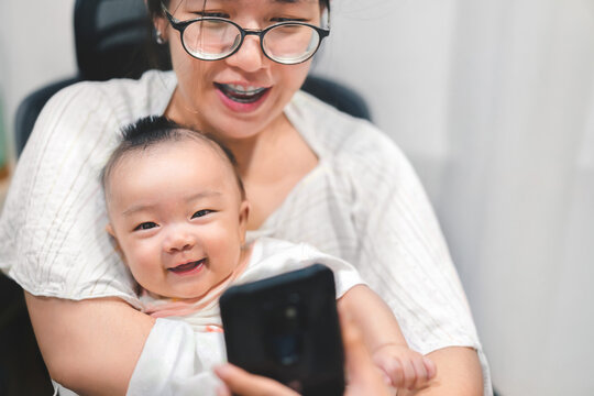 Asian Mother and baby use smartphone for video call , baby sit on mom and smile together for taking photo.
