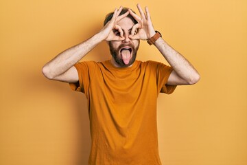 Caucasian man with beard wearing casual yellow t shirt doing ok gesture like binoculars sticking tongue out, eyes looking through fingers. crazy expression.