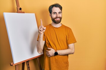 Caucasian man with beard standing by painter easel stand smiling with an idea or question pointing...