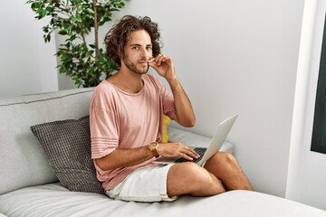 Young hispanic man sitting on the sofa at home using laptop mouth and lips shut as zip with...