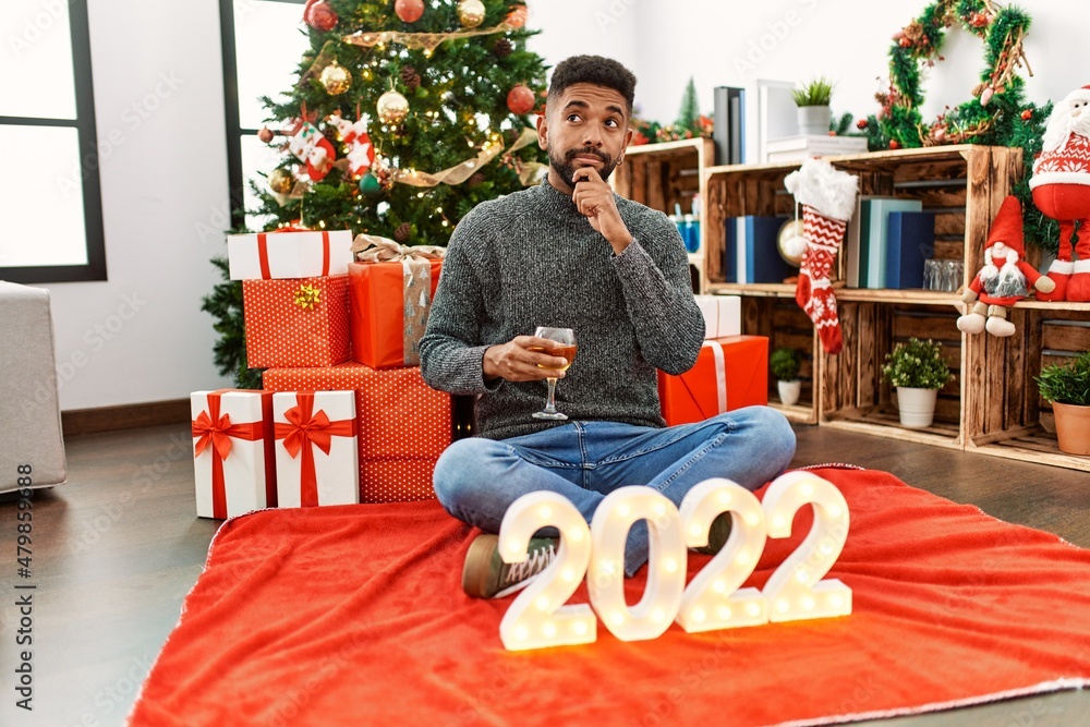 Poster Young hispanic man with beard sitting by christmas tree celebrating 2022 new year serious face thinking about question with hand on chin, thoughtful about confusing idea