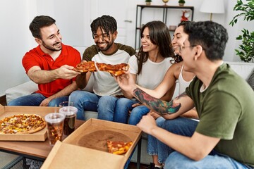 Group of young friends smiling happy eating italian pizza sitting on the sofa at home.