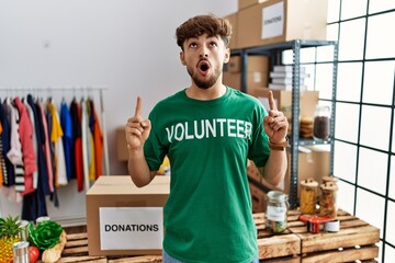 Young arab man wearing volunteer t shirt at donations stand amazed and surprised looking up and pointing with fingers and raised arms.