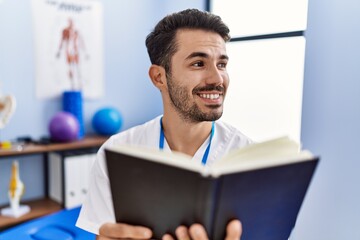 Young hispanic man wearing physiotherapist uniform reading book at rehab clinic