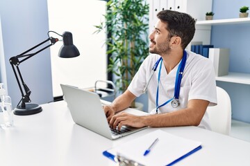 Obraz premium Young hispanic man wearing doctor uniform using laptop working at clinic