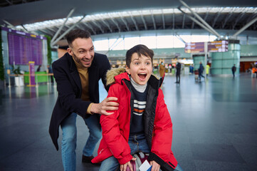 Cheerful happy father rides his son on a luggage in the departure hall of the international airport. Dad and son having fun together while waiting for passport and customs control and boarding