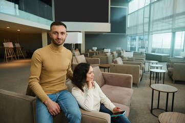 Handsome attractive Caucasian man looks at camera hugging his girlfriend, sitting in chair in an international airport lounge, waiting to board a flight. Multi ethnic couple travels on their honeymoon