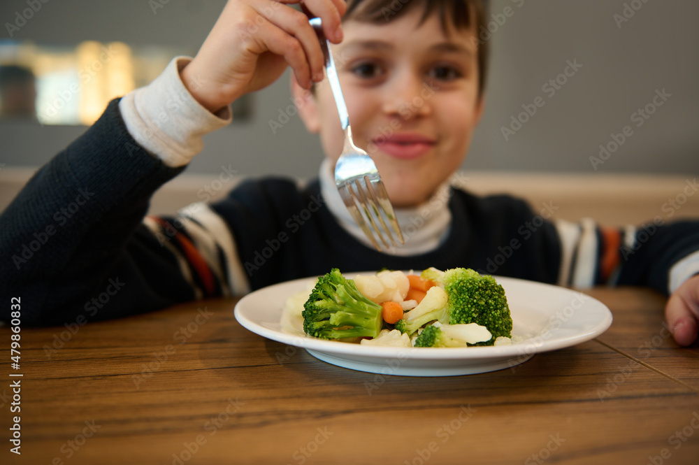 Wall mural Image focused on a plate of steamed vegetables. Adorable smiling Caucasian teenage boy sitting at table and eating healthy lunch of steamed carrot and broccoli. Nutrition, vegetarian diet for toddler