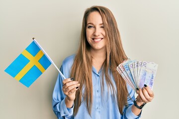 Young blonde woman holding sweden flag and krone banknotes winking looking at the camera with sexy...