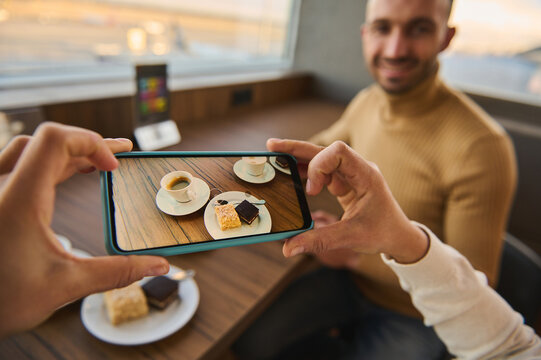 Female hands taking photo on smartphone of cup of coffee and cakes on wooden table with blurred handsome man sitting at panoramic windows overlooking the planes on runways. Mobile phone live view mode