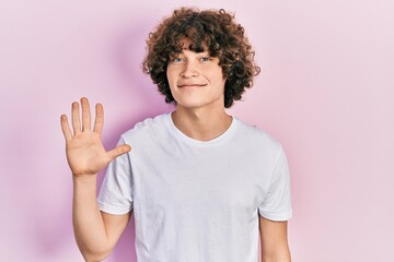 Handsome young man wearing casual white t shirt showing and pointing up with fingers number five while smiling confident and happy.