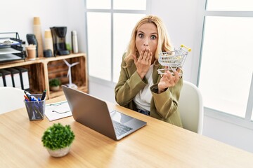 Beautiful blonde woman doing online shopping holding supermarket cart at the office covering mouth...